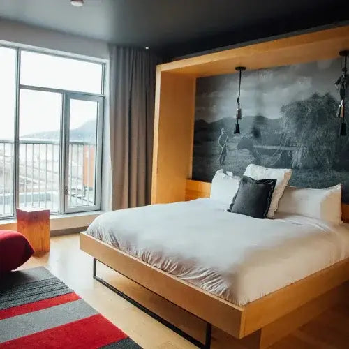 A modern hotel room featuring a minimalist wooden bed frame with crisp white bedding and a dark accent pillow. Behind the bed is a large black-and-white mural depicting a rural hayfield scene. The room has floor-to-ceiling windows with a mountain view, natural light, and a red and grey striped rug adding a pop of colour