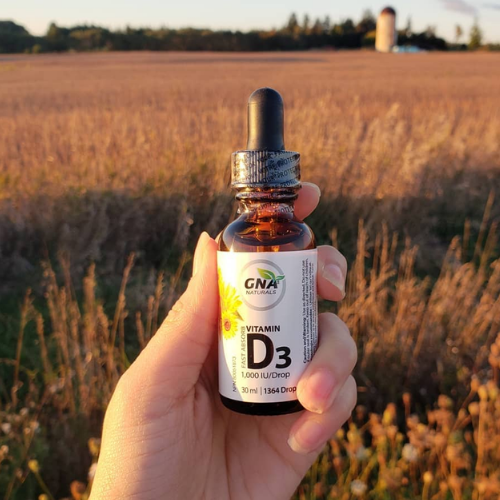 Hand holding a bottle of GNA Naturals Vitamin D3 supplement in front of a field.