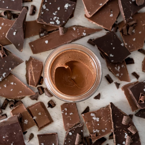 Jar of chocolate pudding surrounded by chocolate pieces on a light background