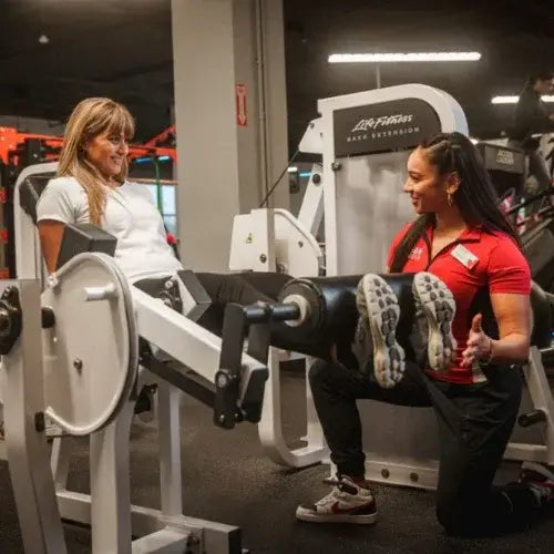 A personal trainer kneeling beside a client who is using a leg extension machine in a gym. The trainer, wearing a red shirt and black pants, offers encouragement while the client, in a white shirt, smiles during the workout. The setting is well-lit with various fitness equipment in the background.