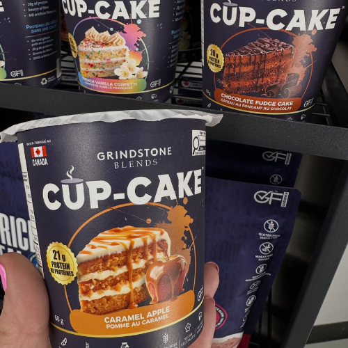 A hand holds a canister of Grindstone Blends Cup-Cake Caramel Apple protein powder in front of other Grindstone Cup-Cake containers on a store shelf.