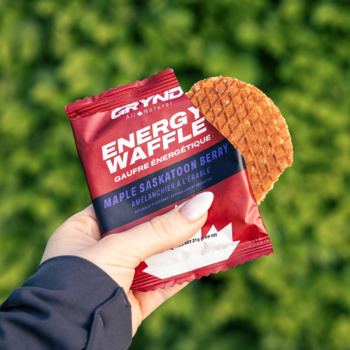 Hand holding a red Grynd energy waffle package with a waffle against a green blurred background
