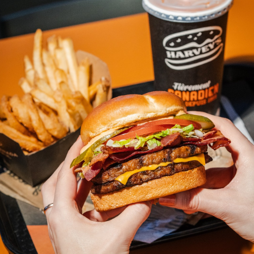Hamburger with cheese, lettuce, and tomato held by hands with fries and a drink in the background.