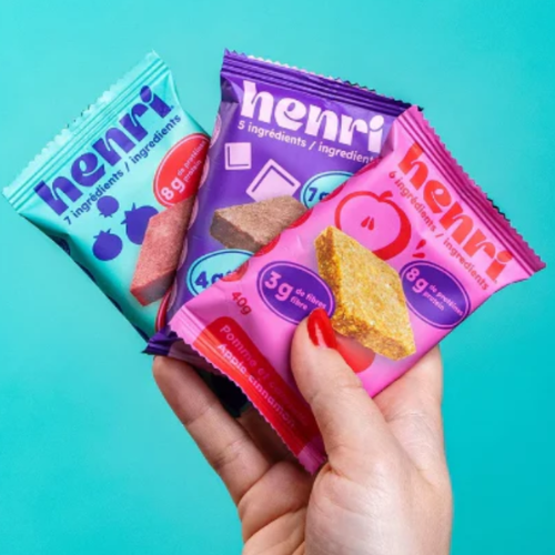 Hand holding three colourful 'henri' snack packages against a turquoise background