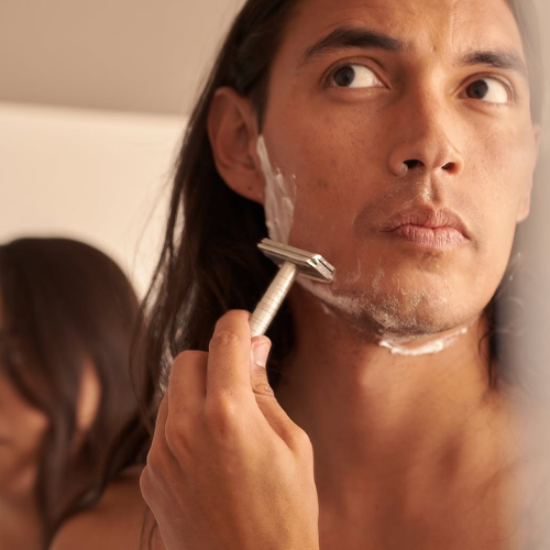 A close-up lifestyle shot of a man with long dark hair shaving his face with a silver safety razor, with shaving cream visible on his cheek. The background is softly blurred, showing another person out of focus.