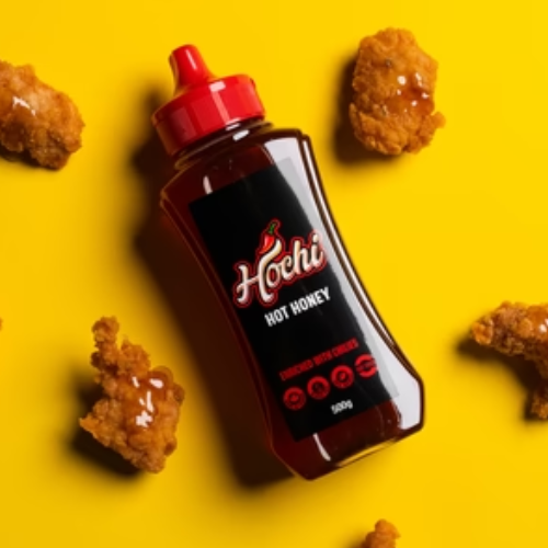 A squeeze bottle of Hochi Hot Honey sauce with a red cap and black label, surrounded by golden fried chicken pieces on a bright yellow background.