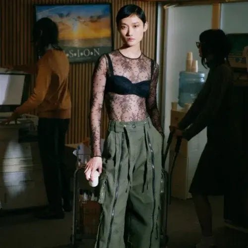 Person wearing a sheer top and green pants in an indoor setting with other people and objects around.