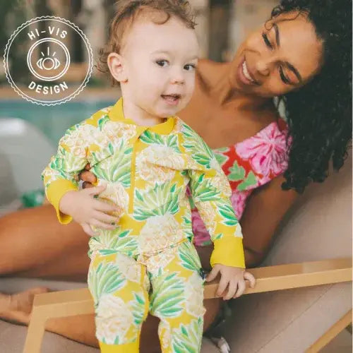 Child wearing a yellow pineapple-patterned Honeysuckle Swim Co. outfit with a woman in a pink swimsuit, both sitting on a deck.
