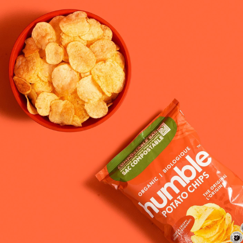 A red bowl filled with potato chips sits on a bright orange background next to a bag of Humble Potato Chips. The bag is orange with green and white branding and shows the text 'Organic Potato Chips' and 'The Original Humble'.
