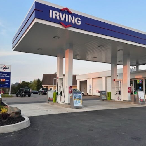 An Irving gas station canopy with multiple fuel pumps, a convenience store building, and a parking lot under a blue sky.
