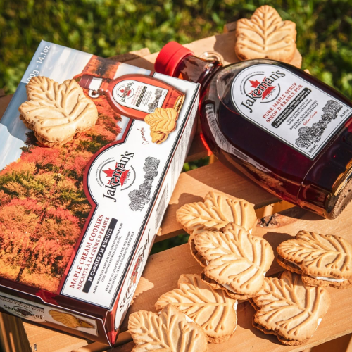 Bottle of maple syrup and box of maple cream cookies on a wooden surface with greenery in the background.