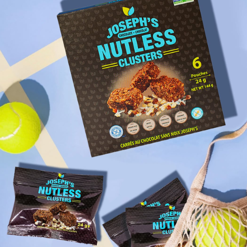 A box and several individually wrapped pouches of Joseph's Nutless Clusters chocolate clusters arranged on a light blue surface with tennis ball props and a mesh bag.