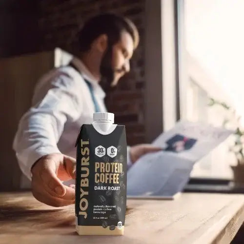 Person holding a box of Joyburst Protein Coffee on a table with a blurred background