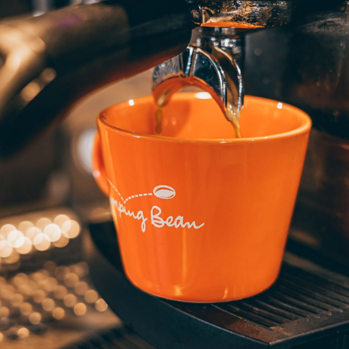 An orange ceramic coffee cup with white 'jumping Bean' script and a coffee bean graphic, filled with espresso under a metal portafilter on a dark espresso machine.