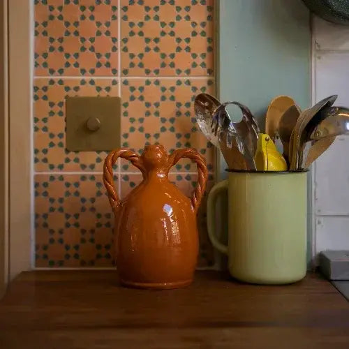 Orange ceramic jug and green mug with utensils on a kitchen counter against a patterned wall.