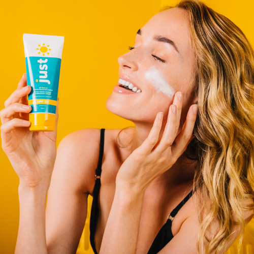 Woman applying sunscreen cream to her face with a 'just' tube against a yellow background