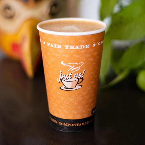 Orange compostable coffee cup with 'just us!' coffee branding on a dark surface.
