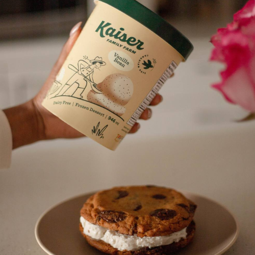 Hand holding a container of Kaiser Family Farm vanilla bean ice cream next to a cookie sandwich on a plate.