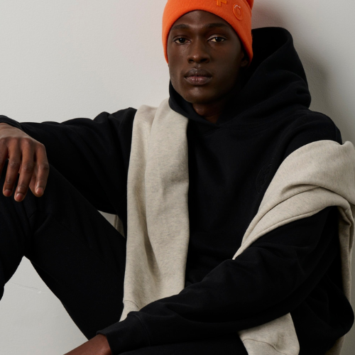 Person wearing a black hoodie, orange beanie, and beige scarf against a white background