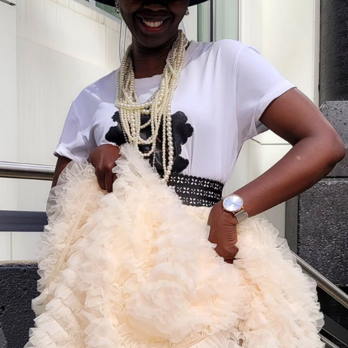 Woman holding a large cream-coloured skirt up, wearing a white top with black floral design and pearl necklace.