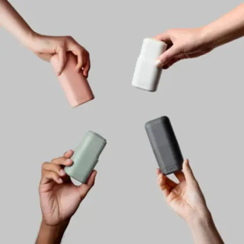 Four hands holding cylindrical deodorant containers in pink, white, green, and gray against a plain background