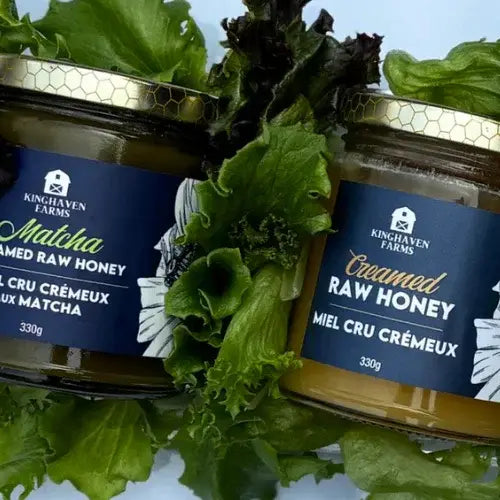 Two jars of Kinghaven Farms creamed raw honey surrounded by green leaves on a light blue background.