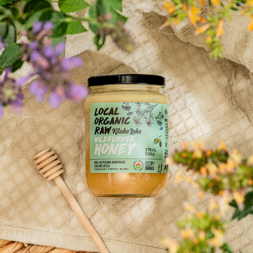 Jar of Local Organic Raw Wildflower Honey with a honey dipper on a textured surface surrounded by flowers.