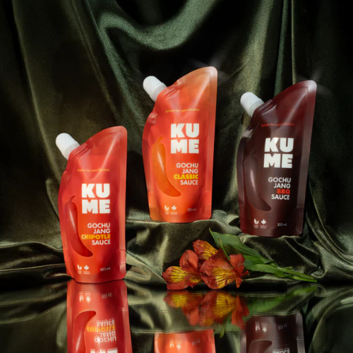 Three bottles of KUME gochujang sauce on a reflective surface with a green fabric background.