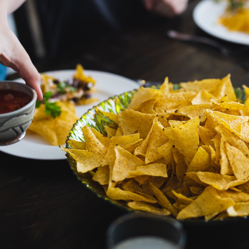 A large green-patterned bowl filled with golden tortilla chips on a dark wooden table, with a hand holding a small bowl of salsa in the foreground and plates of food blurred in the background.