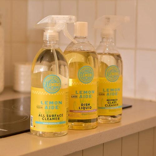 Three bottles of Lemon Aide surface cleaners arranged on a kitchen counter: All Surface Cleaner, Dish Liquid, and Non-Acid Glass Cleaner. The bottles contain a yellow liquid and feature yellow labels with teal text and logos.
