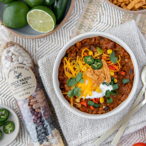  bowl of chili with toppings on a textured surface with limes and a package of Chuckwagon Chili Mix.