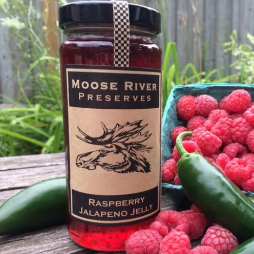 Jar of Moose River Preserves Raspberry Jalapeno Jelly with raspberries and jalapenos on a wooden surface.