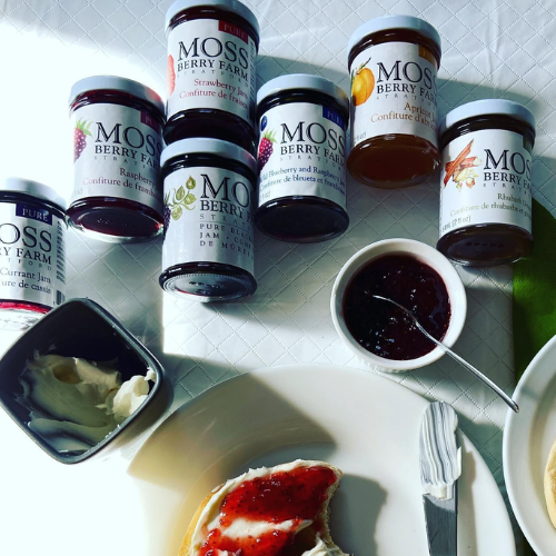 Jars of Moss Berry Farm jam on a table with a bowl of jam and a slice of cake.