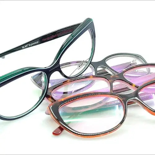 A close-up display of stylish, colourful eyeglass frames arranged in a scattered stack. The glasses come in various cat-eye and rounded shapes with vibrant hues including teal, purple, and tortoiseshell. The inside of one arm reads 'Handcrafted in Colombia' and 'N49 Eyewear.'