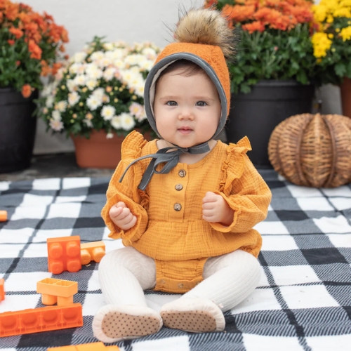 Baby in a yellow outfit and orange hat sitting on a checkered blanket with pumpkins and flowers in the background.