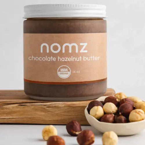 Jar of nomz chocolate hazelnut butter with a bowl of hazelnuts on a wooden board.