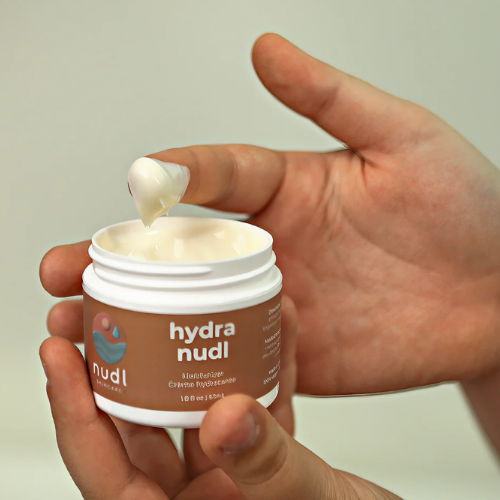 Hand holding a jar of 'hydra nudl' cream with a small amount being applied.
