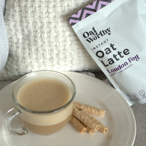 Glass of oat latte with wafer cookies on a white plate, next to a package of Oat Worthy Instant Oat Latte London Fog.