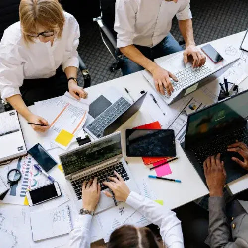 Top-down view of a busy office meeting with several people working around a cluttered table filled with laptops, tablets, smartphones, papers, pens, and charts. The group appears to be collaborating on business or marketing tasks.