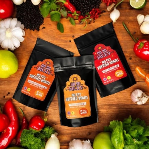 Three packages of 'Oh So Mexy' no fry refried beans on a wooden surface with vegetables.