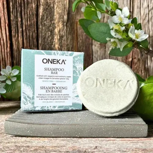 A natural shampoo bar from ONEKA displayed on a stone slab alongside its eco-friendly box packaging. The label highlights organic ingredients like white pine and geranium. Fresh greenery and white blossoms decorate the rustic wooden background, emphasizing a clean and botanical aesthetic.
