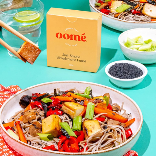 Bowl of noodles with vegetables, oomé tofu box, and glasses of water on a teal background