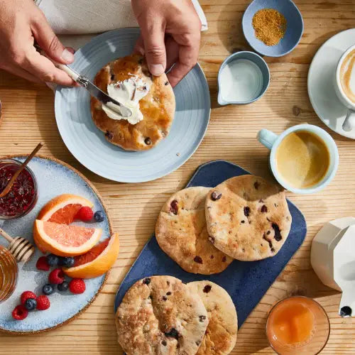 Person spreading butter on a pancake with a breakfast setting including coffee, fruit, and pastries.