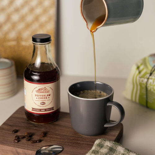 Bottle of maple syrup being poured into a mug with a wooden cutting board and coffee beans on a surface.