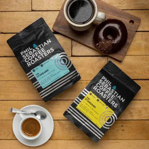 Two bags of Phil & Sebastian Coffee Roasters on a wooden table with a cup of coffee and a donut.