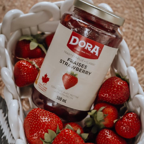 Jar of Dora strawberry preserves surrounded by fresh strawberries in a white basket.