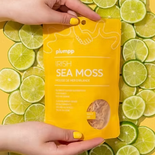 A person's hands holding a yellow pouch of 'plumpp' Irish Sea Moss, set against a background of sliced lime green rinds.