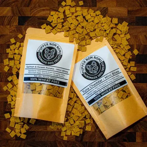 Two packages of Pupper Nom Noms handmade dog treats on a wooden surface with treats scattered around.