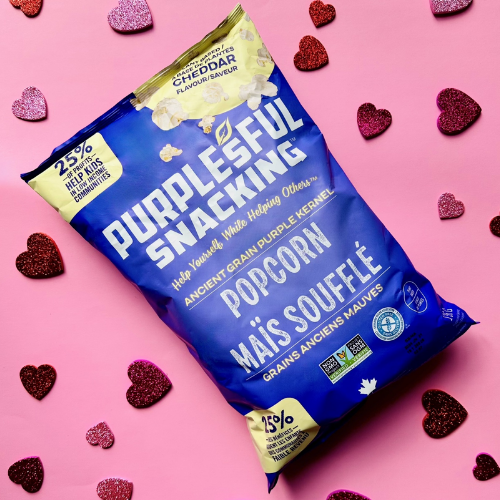 Blue bag of Purplesful Snacking popcorn on a pink background with decorative hearts.