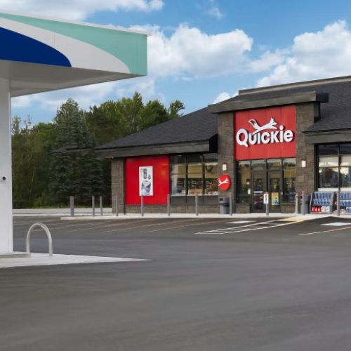Quickie convenience store exterior with a red and white sign, dark roof, stone facade, and a gas station canopy in the foreground under a blue sky with clouds.